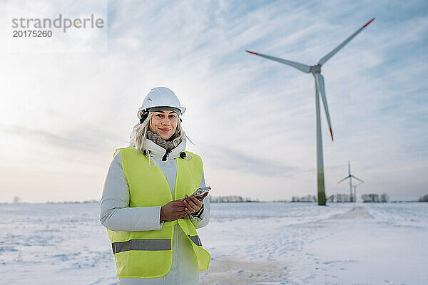 Lächelnde Frau mit Mobiltelefon steht am Windpark