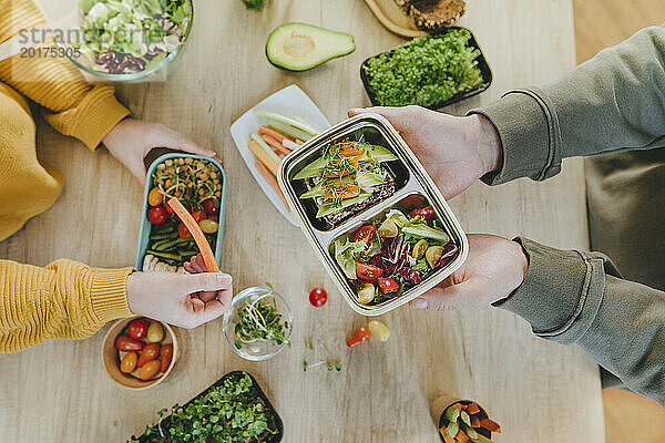 Hände eines Mannes  der eine vegetarische Lunchbox neben einer Frau am Tisch hält