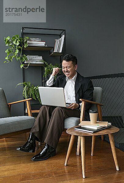 Erfolgreicher Geschäftsmann jubelt und sitzt mit Laptop im Sessel
