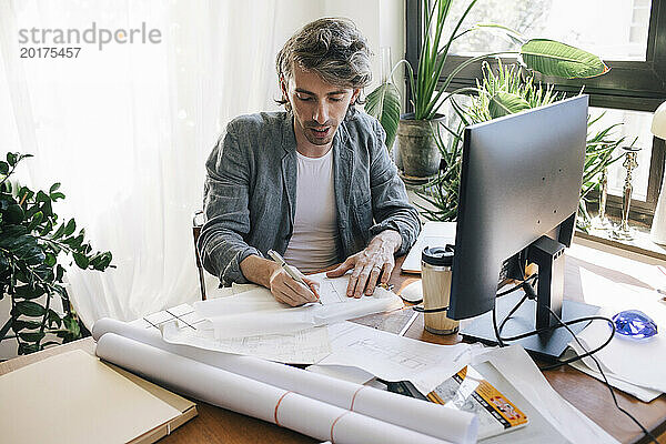 Selbstbewusster männlicher Architekt skizziert am Schreibtisch im Heimbüro einen Bauplan auf Papier