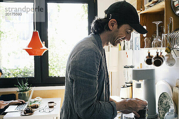 Seitenansicht eines lächelnden männlichen Architekten  der Kaffee zubereitet  während er in der Küche im Heimbüro steht
