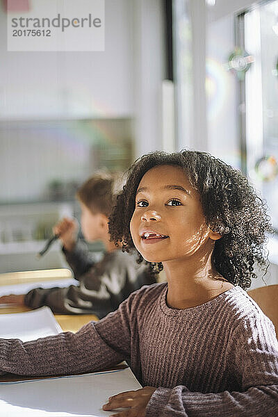 Lächelndes Mädchen mit lockigem Haar  das wegschaut  während es im Klassenzimmer im Kindergarten sitzt