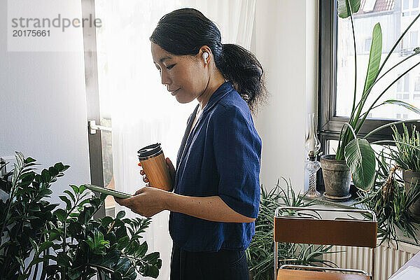 Seitenansicht einer Architektin mit wiederverwendbarer Kaffeetasse und Smartphone im Heimbüro