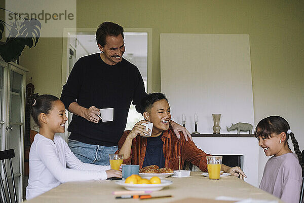 Glückliche schwule Eltern genießen das Frühstück mit ihren Töchtern  während sie zu Hause am Esstisch sitzen