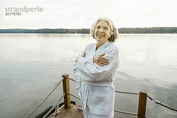 Porträt einer glücklichen Seniorin im weißen Bademantel  die sich auf einem Hausboot am Fluss vor dem Himmel umarmt