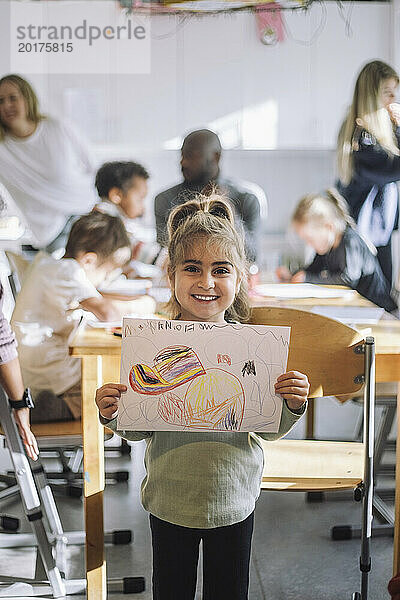 Porträt eines lächelnden Mädchens  das Zeichnung zeigt  während es neben einer Bank im Klassenzimmer im Kindergarten steht