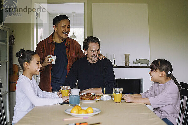 Glückliches schwules Paar  das Spaß beim Frühstück mit Töchtern am Esstisch zu Hause hat