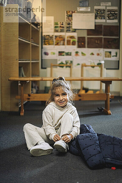 Porträt eines lächelnden Mädchens  das Schnürsenkel bindet  während es im Klassenzimmer im Kindergarten auf dem Boden sitzt