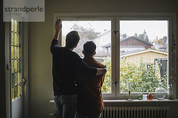 Rückansicht eines Mannes mit einem Arm um ein schwules Paar  das zu Hause durch das Fenster schaut