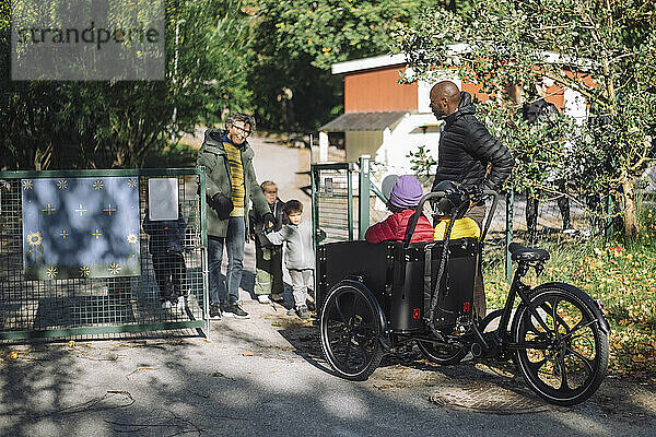 Lehrerin steht am Tor und begrüßt Kinder  die im Lastenfahrrad im Kindergarten sitzen