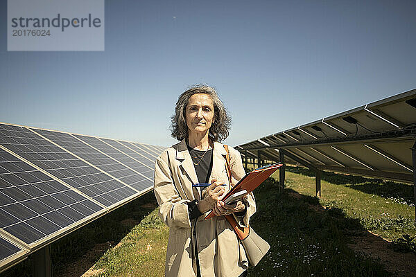 Porträt einer selbstbewussten älteren Unternehmerin  die einen Ordner in der Hand hält  während sie in der Nähe von Solarpaneelen auf dem Feld steht