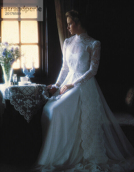 Junge Frau im langen weißen Brautkleid sitzt drinnen am Fenster.