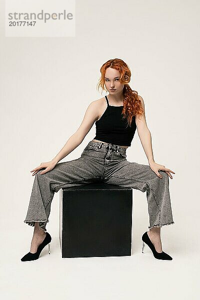 Selbstbewusstes junges weibliches Model mit lockigem rotem Haar in trendigem Outfit sitzt breitbeinig auf einem Stuhl und schaut in die Kamera