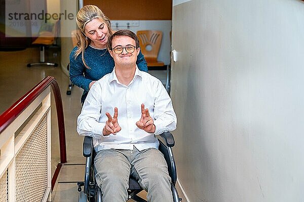 Gesundheitsarbeiter  der einen glücklichen Mann mit Down Syndrom im Rollstuhl schiebt  während er mit den Fingern erfolgreich gestikuliert