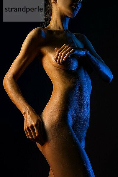 Sinnliche nackte weibliche Modell bedeckt Brust  während in dunklen Studio in Farbe Licht gegen schwarzen Hintergrund stehen