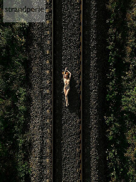 Draufsicht auf eine nackte Frau im Liegen auf einer Bahnstrecke bei Sonnenaufgang