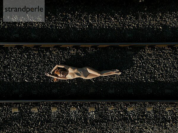 Draufsicht auf eine nackte Frau im Liegen auf einer Bahnstrecke bei Sonnenaufgang