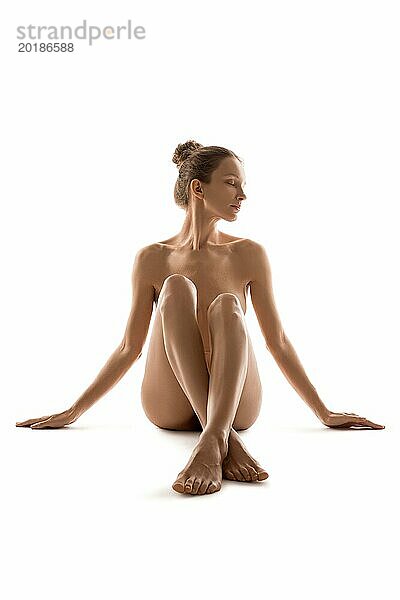 Ganzkörper von zarten nackten weiblichen Modell mit geschlossenen Augen bedeckt nackten Brust mit gekreuzten Beinen im Studio gegen weißen Hintergrund