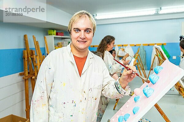 Porträt eines geistig behinderten Mannes in weißem Laborkittel  der in der Malklasse in die Kamera lächelt