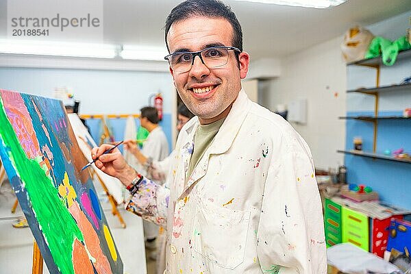 Behinderter Mann schaut in die Kamera und lächelt glücklich beim Malen auf Leinwand