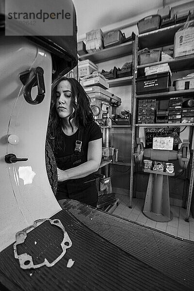 Ernsthafte Mechanikerin  die ein Teil an einem Moped zerlegt  italienischer Oldtimer Motorroller in der Werkstatt  echte Frauen  die traditionelle Männerarbeiten der Vergangenheit ausführen  Schwarz Weiß Foto