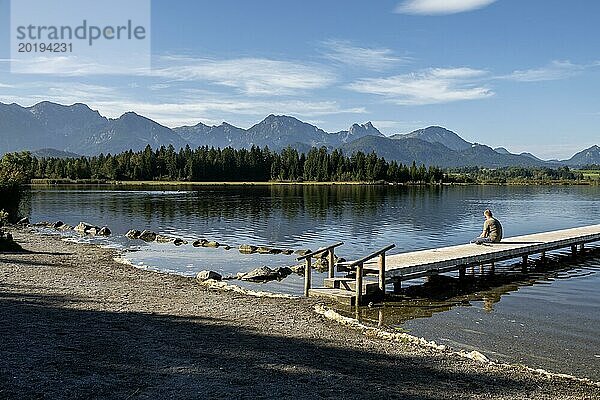 Person sitzt auf einem Holzsteg und blickt auf einen ruhigen See mit Bergen im Hintergrund  Hopfensee  Hopfen am See  bei Füssen  Allgäuer Alpen  Ostallgäu  Allgäu  Bayern  Deutschland  Europa