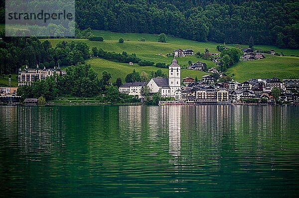 Idyllische Dorfansicht mit Kirche am Ufer eines ruhigen Sees  umgeben von grünen Wiesen und Wäldern  Sankt Wolfgang  Wolfgangsee  Salzkammergut  Östereich