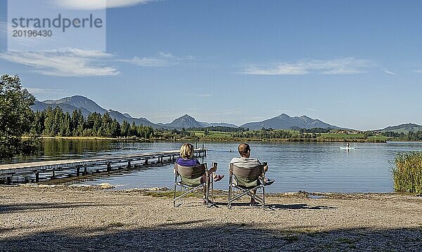 Zwei Menschen sitzen auf Stühlen am Seeufer und blicken auf eine malerische Berglandschaft  Hopfensee  Hopfen am See  bei Füssen  Allgäuer Alpen  Ostallgäu  Allgäu  Bayern  Deutschland  Europa