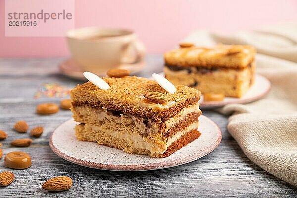 Honigkuchen mit Milchcreme  Karamell  Mandeln und einer Tasse Kaffee auf grauem und rosa Hintergrund und Leinenstoff. Seitenansicht  Nahaufnahme  selektiver Fokus