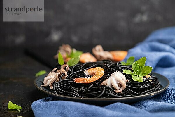 Schwarze Tintenfischnudeln mit Garnelen und kleinen Tintenfischen auf schwarzem Betonhintergrund und blauem Textil. Seitenansicht  Nahaufnahme  selektiver Fokus