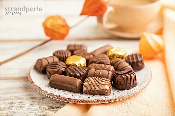 Schokoladenbonbons mit Kaffeetasse und Physalisblüten auf weißem Holzhintergrund und orangefarbenem Textil. Seitenansicht  Nahaufnahme  selektiver Fokus