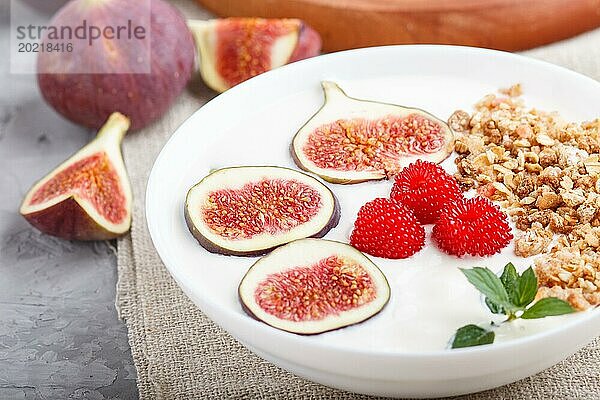 Joghurt mit Himbeeren  Müsli und Feigen in einem weißen Teller auf einem grauen Betonhintergrund und Leinentextil. Seitenansicht  Nahaufnahme  selektiver Fokus