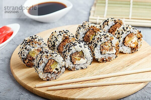 Japanische Maki Sushi Rollen mit Lachs  Sesam  Gurken Essstäbchen  Sojasauce und mariniertem Ingwer auf einem Holzbrett vor einem grauen Betonhintergrund. Seitenansicht  Nahaufnahme  selektiver Fokus