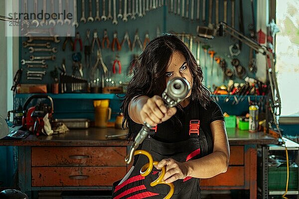Intensive sexy Frau Mechanikerin zielt ein Werkzeug auf die Kamera in einer Werkstatt  eine komplette Werkzeugtafel im Hintergrund mit Bokeh Effekt  traditionelle männliche Arbeitsplätze von Mixed race latino Frau