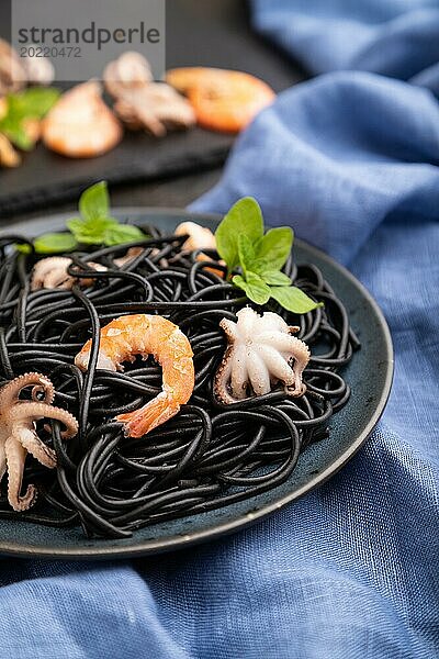 Schwarze Tintenfischnudeln mit Garnelen und kleinen Tintenfischen auf schwarzem Betonhintergrund und blauem Textil. Seitenansicht  Nahaufnahme  selektiver Fokus