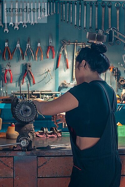 Mechanikerin  die eine Lichtmaschine an einer Werkbank in einer Autowerkstatt repariert  trägt einen schwarzen Overall und ein schwarzes Hemd  eine komplette Werkzeugtafel im Hintergrund mit Bokeh Effekt  traditionelle Männerberufe von Mixed race latino woman