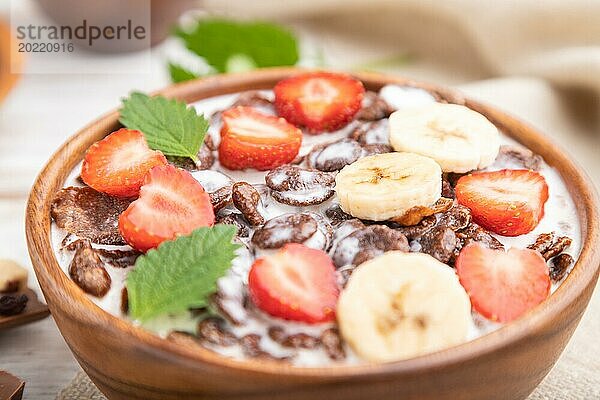 Schokoladen Cornflakes mit Milch und Erdbeeren in einer Holzschale auf weißem Holzhintergrund und Leinenstoff. Seitenansicht  Nahaufnahme  selektiver Fokus