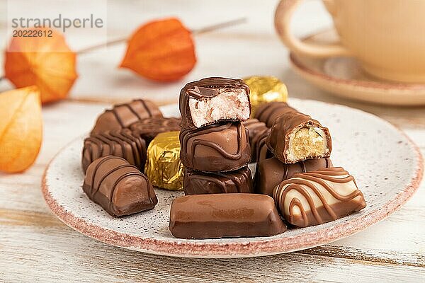 Schokoladenbonbons mit Kaffeetasse und Physalisblüten auf weißem Holzhintergrund und orangefarbenem Textil. Seitenansicht  Nahaufnahme  selektiver Fokus