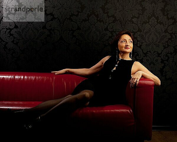 Schönheit reife Frau sitzen auf rotem Sofa im dunklen Raum