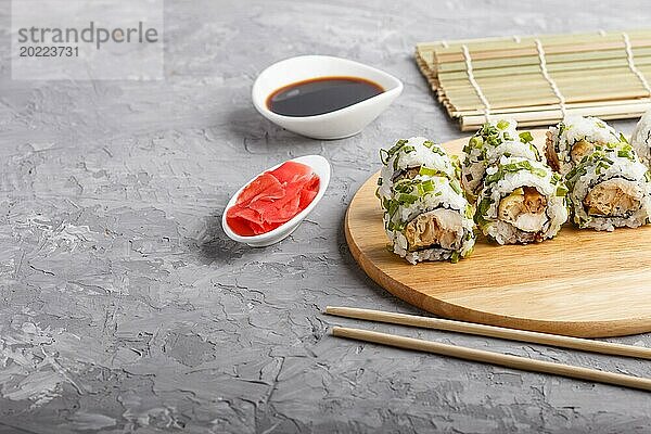 Japanische Maki Sushi Rollen mit grünen Zwiebeln  Essstäbchen  Sojasauce und mariniertem Ingwer auf einem Holzbrett vor einem grauen Betonhintergrund. Seitenansicht  Kopierraum  selektiver Fokus