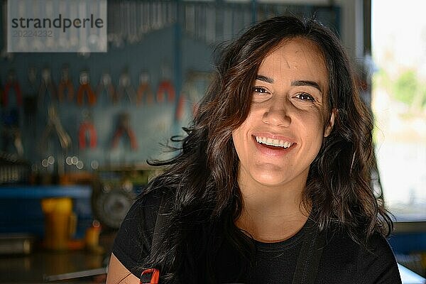 Lächelnde hispanische Mechanikerin in der Werkstatt vor einer Werkbank stehend  eine komplette Werkzeugtafel im Hintergrund mit Bokeh Effekt  traditionelle Männerberufe von Mixed race latino woman