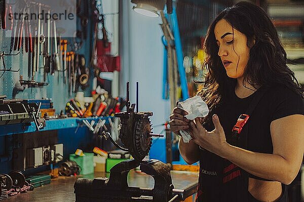 Ein weiblicher Mechaniker in einem Overall in einer Werkstatt reinigt sorgfältig Motorradteile  eine komplette Werkzeugtafel im Hintergrund mit Bokeh Effekt  traditionelle männliche Berufe von Mixed race latino woman