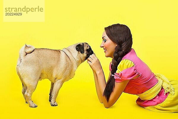Glückliches Mädchen wie niedliche Puppe füttern einen kleinen Hund auf gelbem Hintergrund
