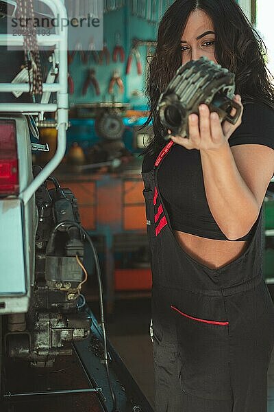 Professionelle latino weiblichen Motorrad Mechaniker hält Komponente der italienischenvintage Roller in einer Werkstatt  ein komplettes Werkzeug Panel im Hintergrund mit Bokeh Effekt  traditionelle männliche Arbeitsplätze von Mixed race hispanische Frau