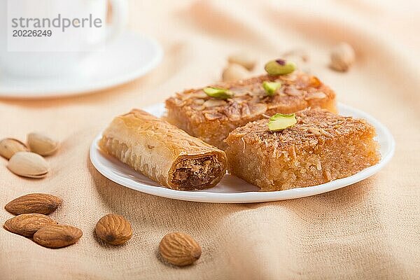 Traditionelle arabische Süßigkeiten (Basbus  Kunafa  Baklava) und eine Tasse Kaffee auf einem grauen Betonhintergrund und orangefarbenem Textil. Seitenansicht  Nahaufnahme  selektiver Fokus  Makro