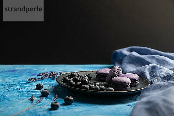 Lila Macarons oder Makronen Kuchen mit Blaubeeren auf Keramikteller auf einem blauen und schwarzen Hintergrund und blauem Textil. Seitenansicht  Kopierraum  selektiver Fokus