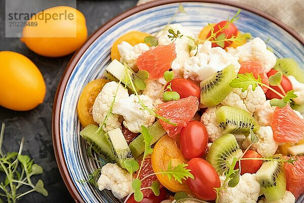 Vegetarischer Salat aus Blumenkohl  Kiwi  Tomaten  Mikrogrünsprossen auf schwarzem Betonhintergrund und Leinentuch. Seitenansicht  Nahaufnahme  selektiver Fokus