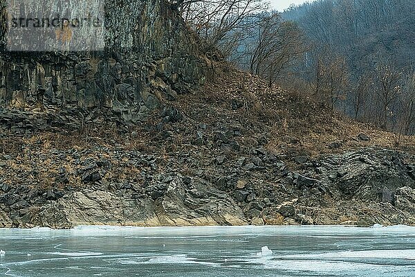 Wand aus vulkanischem Gestein am Ufer eines zugefrorenen Flusses mit Hintergrund aus dichten Bäumen an einem Wintertag in der Provinz Gyeonggi in Südkorea