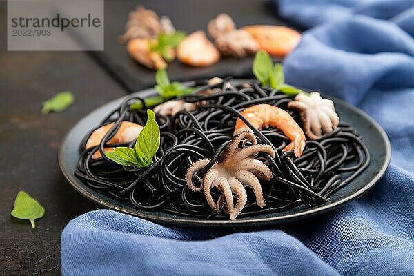 Schwarze Tintenfischnudeln mit Garnelen und kleinen Tintenfischen auf schwarzem Betonhintergrund und blauem Textil. Seitenansicht  Nahaufnahme  selektiver Fokus