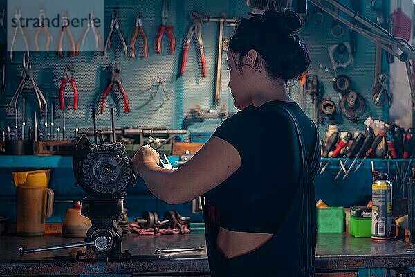 Geschickte Handwerkerin bei der Reparatur eines Motorradmotorteils in einer Werkstatt mit blaugrüner Wand  eine komplette Werkzeugtafel im Hintergrund mit Bokeh Effekt  traditionelle Männerberufe von Mixed race latino woman